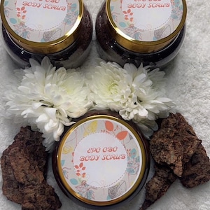 Puede incluir: Tres frascos de vidrio con tapas doradas llenos de un exfoliante marrón oscuro. Cada frasco tiene una etiqueta blanca con el texto "EPO OBO BODY SCRUB" en letras negras. Los frascos están rodeados de flores blancas y corteza marrón.