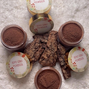 Puede incluir: Una variedad de recipientes llenos de polvo marrón, etiquetados como "EPO OBO POWDER", junto con trozos de corteza de árbol. Los recipientes tienen tapas doradas y diseños florales. Los artículos están dispuestos sobre una superficie blanca texturizada.