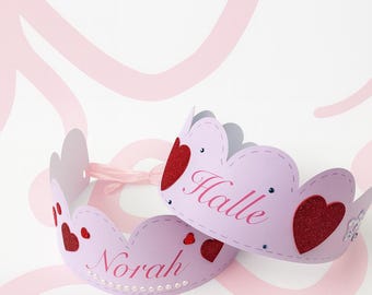 May include: Two lilac paper crowns with heart details and the names "Halle" and "Norah" in pink script. The crowns have a scalloped edge and are joined by a pink ribbon.