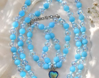 Puede incluir: Un conjunto de joyería compuesto por un collar y una pulsera hechos de cuentas azul claro mate alternadas con cuentas transparentes de purpurina iridiscente. Ambas piezas incluyen un colgante en forma de corazón con un patrón de escamas.