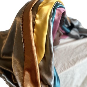 Op de afbeelding: Een set van zes satijnen sjaals in verschillende kleuren, waaronder teal, geel, goud, roze en grijs. De sjaals zijn in lagen gelegd, waardoor een regenboog-effect ontstaat.
