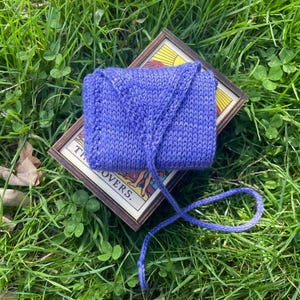 May include: A purple knitted pouch with a drawstring closure, sitting on top of a deck of tarot cards. The cards are in a wooden box with the text "The Lovers."