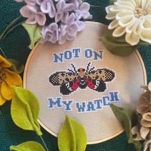 May include: A cross-stitch embroidery hoop with the words "NOT ON MY WATCH" in blue. The design features a detailed insect with black, brown, and red accents. The hoop is surrounded by felt flowers and leaves in yellow, white, and purple.