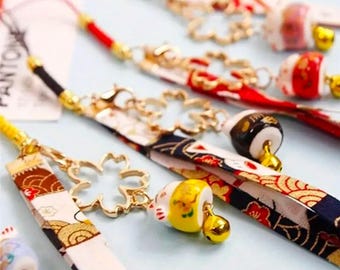May include: Close-up of several Japanese-style keychains. Each keychain features a patterned fabric strap with gold-colored metal accents, charms, and small bells. The keychains have various designs, including floral and cat motifs, with red, white, and blue colors.
