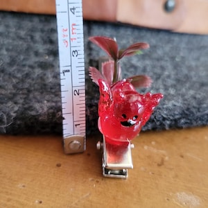 Puede incluir: Pinza para el pelo de resina roja translúcida con forma de personaje de dibujos animados, con una pequeña planta que brota de la parte superior. La pinza mide aproximadamente 2,5 cm de largo y tiene un cierre de metal plateado.