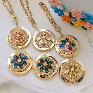 以下が含まれることがあります： 金色の花の模様のバッグハンガーが6つ。各ハンガーは、カラフルな宝石で作られた中央の花と、小さなクリスタルで囲まれた円形のデザインが特徴です。ハンガーはゴールドチェーンに取り付けられています。