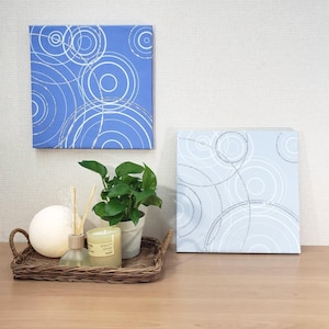 May include: Two square canvas art pieces with a blue background and white circular designs. One is a darker blue, the other a lighter blue. A woven tray holds a white sphere, a candle, a diffuser, and a potted plant.