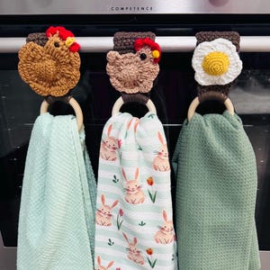 May include: Three kitchen towel holders with crochet toppers. The toppers are shaped like a chicken, a chicken, and a fried egg. Each holder has a wooden ring and a different coloured towel: mint green, bunny print, and sage green.