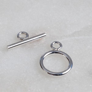 May include: A pair of silver-colored toggle clasps. One clasp is a small, straight bar with a loop on one end. The other is a circular ring with a loop at the top. The clasps are designed for jewelry making.