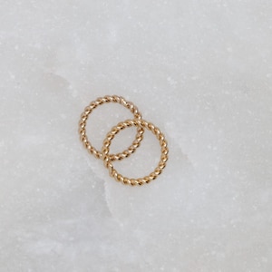 May include: Two gold-colored, twisted-rope style rings are interlocked on a white marble surface. The rings are small and delicate, with a textured appearance. The rings are likely jewelry, possibly earrings or small hoops.