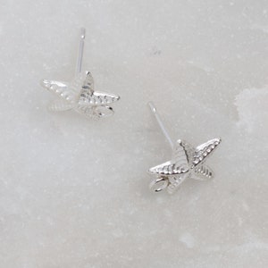 May include: A pair of silver-colored starfish stud earrings. Each earring features a detailed starfish design with textured surfaces and a post for pierced ears. The earrings are displayed on a white marble surface.