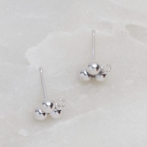 May include: A pair of silver stud earrings with three small balls clustered together. Each earring has a small loop at the top for attaching to a post.