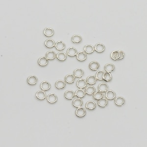 May include: A scattering of small, silver jump rings on a white background. These circular rings are used in jewellery making and crafts. The rings are made of a shiny metal and are all the same size.