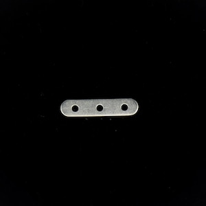 Puede incluir: Una pequeña placa metálica plateada con tres agujeros espaciados uniformemente. La placa es rectangular con extremos redondeados. El objeto se encuentra sobre un fondo negro sólido, resaltando su forma y diseño.