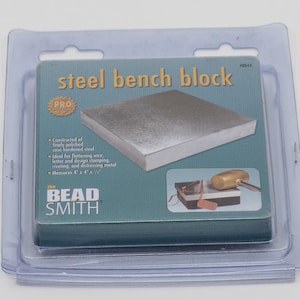 Könnte beinhalten: Eine blaue und grüne Verpackung für einen Stahl-Werkbankblock mit der Aufschrift "steel bench block PRO". Die Verpackung zeigt einen silbernen Metall-Werkbankblock und einen Hammer, der auf ein Stück Metall schlägt. Der Text auf der Verpackung lautet: "Hergestellt aus schwerem Guss, neu poliert, verchromtem gehärtetem Stahl. Ideal zum Glätten von Draht, besser zum Strukturieren, Prägen, Verfolgen und Verformen von Metall. Maße 4" x 6" x 7/8". The Bead Smith."