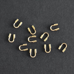 May include: A collection of small, gold-colored U-shaped jewelry findings scattered on a dark gray surface. Each finding has a curved body with two parallel, open ends, designed for connecting jewelry components.