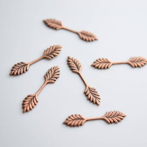 Op de afbeelding: Zes koperkleurige metalen versieringen in de vorm van bladeren. Elk stuk heeft een centrale steel met gedetailleerde bladerpatronen aan beide uiteinden. De versieringen zijn gerangschikt op een wit oppervlak, ideaal voor knutselen of sieraden.