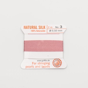 May include: A spool of pink natural silk thread for stringing pearls and beads. The spool is labeled "NATURAL SILK 2 m No. 3 100% Naturseide Ø 0,50 mm" and "www.griffin.de For stringing pearls and beads".