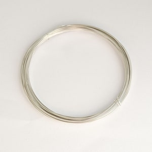 Puede incluir: Una bobina de alambre plateado, enrollada firmemente en forma circular. El alambre parece liso y reflectante, lo que sugiere un material metálico. La imagen está sobre un fondo blanco liso, resaltando la forma y textura del alambre. Probablemente se trate de suministros para joyería o manualidades.