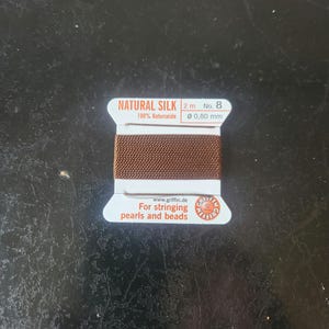 May include: A spool of brown natural silk thread for stringing pearls and beads. The label reads "NATURAL SILK 2 m No. 8 100% Naturseide Ø 0,80 mm www.griffin.de For stringing pearls and beads".