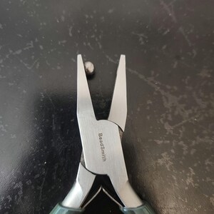 May include: Close-up of a pair of silver beading pliers with green handles. The pliers are open and the text "BeadSmith" is visible on the metal.