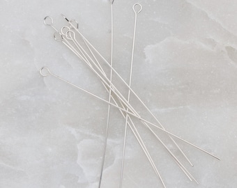 May include: A set of silver-toned head pins with a loop at the top. The pins are arranged in a fan shape on a white marble surface.