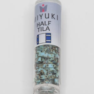 Op de afbeelding: Een doorzichtige plastic buis gevuld met lichtblauwe en bruine Miyuki Half Tila kralen. De buis heeft een label met de tekst "MIYUKI HALF TILA" in blauw en wit. De kralen zijn klein, rechthoekig en hebben een verweerde uitstraling.
