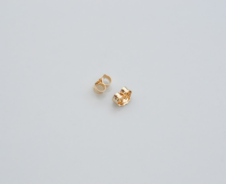 K&ouml;nnte beinhalten: Zwei goldene Ohrringverschl&uuml;sse, einer ist ein Standard-Druckverschluss und der andere ein Schmetterlingsverschluss.
