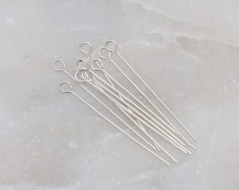 May include: A group of silver head pins with a loop at the top. The pins are arranged in a fan shape on a white marble surface.
