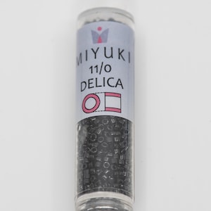 Peut inclure: Un petit flacon en plastique transparent rempli de perles de rocaille noires. L'étiquette sur le flacon indique "MIYUKI 11/0 DELICA" avec un logo de couronne au-dessus du texte. L'étiquette comprend également un cercle et un carré.