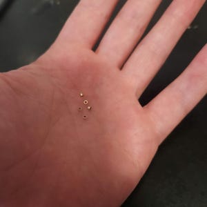 May include: A close-up of a hand holding six small, gold-coloured jump rings.