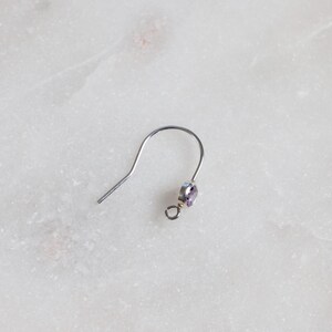 May include: A single silver earring hook with a small, round, pale purple gemstone. The earring hook is curved and has a simple, minimalist design. The gemstone is set in a silver bezel.