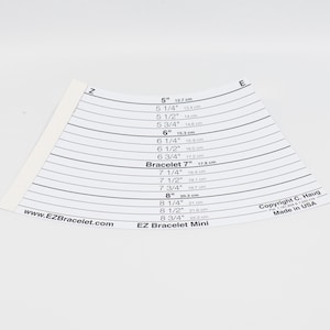 May include: A white plastic bracelet sizing tool with a curved shape and measurements in inches and centimetres. The tool is labelled "EZ Bracelet Mini" and includes sizes from 5 inches to 8 3/4 inches.