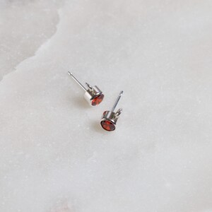 May include: A pair of stud earrings with a silver setting and a round, deep red gemstone. The earrings are displayed on a white, marble-like surface. The earrings are a classic jewelry item.