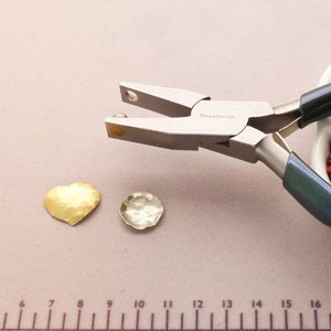 May include: Close-up of a silver jewelry pliers with a round head, used to crimp metal findings. Two gold-colored metal heart-shaped findings are shown on a beige surface with a ruler.