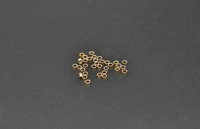 May include: A group of small, gold-coloured jump rings scattered on a dark grey surface.