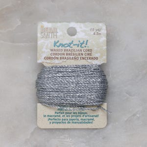 May include: A spool of silver Knot-it! waxed Brazilian cord, 15 yards (4.5 metres) long. The label reads "Knot-it! Waxed Brazilian Cord, Cordon Brésilien Ciré, Cordón Brasileño Encerado". It is perfect for jewellery, macrame, and craft projects.