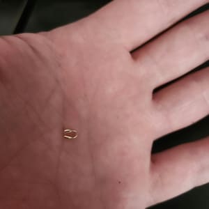 May include: A small, gold-colored metal clasp, shaped like a curved staple, rests in the palm of a hand. The clasp appears to be a jewelry finding or a component for a small craft project. The clasp is a simple, minimalist design.