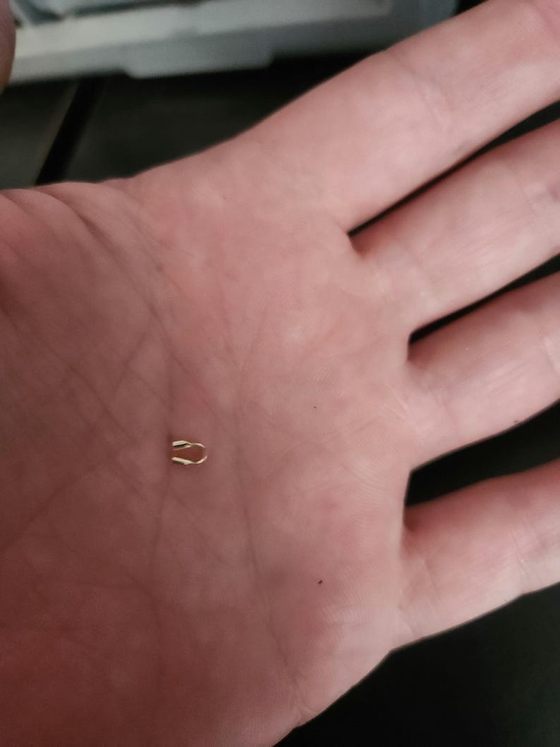 May include: A small, gold-colored metal clasp, shaped like a curved staple, rests in the palm of a hand. The clasp appears to be a jewelry finding or a component for a small craft project. The clasp is a simple, minimalist design.