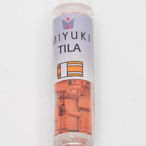 Può includere: Un piccolo flacone di plastica trasparente riempito con perline di piastrelle arancioni. L'etichetta sul flacone riporta "MIYUKI TILA" con un logo a corona sopra il testo. L'etichetta include anche una piccola grafica di una perlina di piastrella.