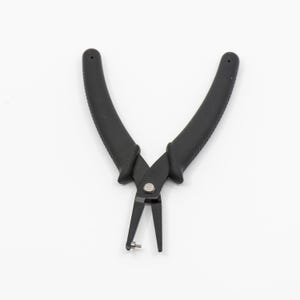 May include: Black jewelry pliers with curved, textured handles and pointed tips. The pliers are open, revealing a small metal pin at the tip. The tool is designed for precision work in jewelry making and crafts.