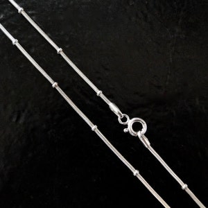 Puede incluir: Dos cadenas de serpiente plateadas con peque&ntilde;as cuentas, una es m&aacute;s larga que la otra. Las cadenas est&aacute;n sobre un fondo negro.