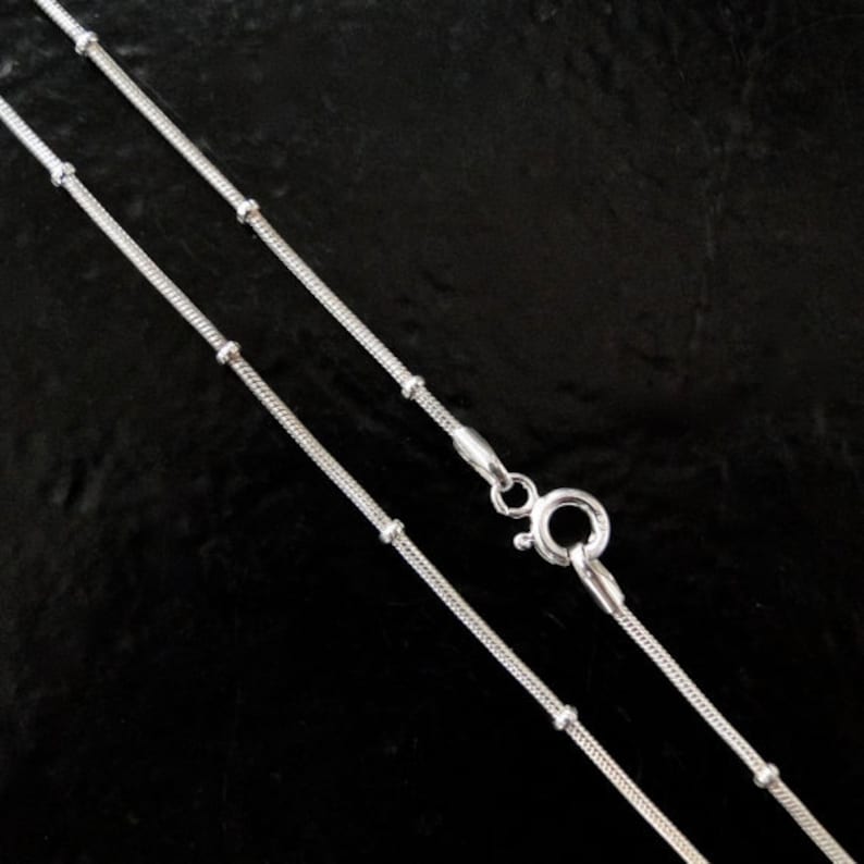 Puede incluir: Dos cadenas de serpiente plateadas con peque&ntilde;as cuentas, una es m&aacute;s larga que la otra. Las cadenas est&aacute;n sobre un fondo negro.
