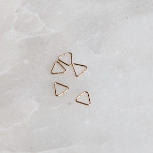 May include: Six gold triangle-shaped metal rings. The rings are small and delicate, and they are arranged in a loose cluster on a white surface.
