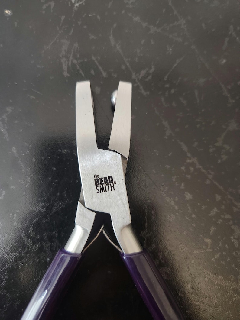 K&ouml;nnte beinhalten: Nahaufnahme einer silbernen Perlen-Zange mit violetten Griffen. Die Zange ist mit "the Bead Smith" beschriftet.