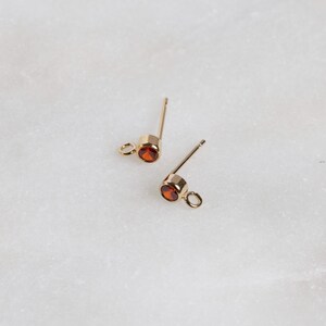 May include: A pair of gold-toned earring posts with small red gemstone accents.