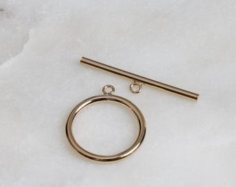 May include: Gold-toned metal toggle clasp with a round ring and a straight bar. The clasp is used for necklaces, bracelets, or other jewelry.
