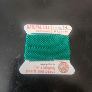 May include: A spool of green natural silk thread for stringing pearls and beads. The label reads "NATURAL SILK 2 m No. 14 100% Naturseide Ø 1,02 mm www.griffin.de For stringing pearls and beads"