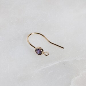 Könnte beinhalten: Ein goldener Ohrring mit einem violetten Edelstein. Der Ohrring ist ein einfacher Haken-Stil mit einer kleinen Schlaufe am Ende.