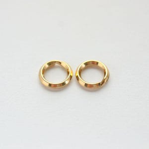 May include: Two small, circular gold-toned metal jump rings. Each ring features a smooth, polished surface with a visible split opening on one side, designed for connecting jewelry components or other small items in crafting projects.